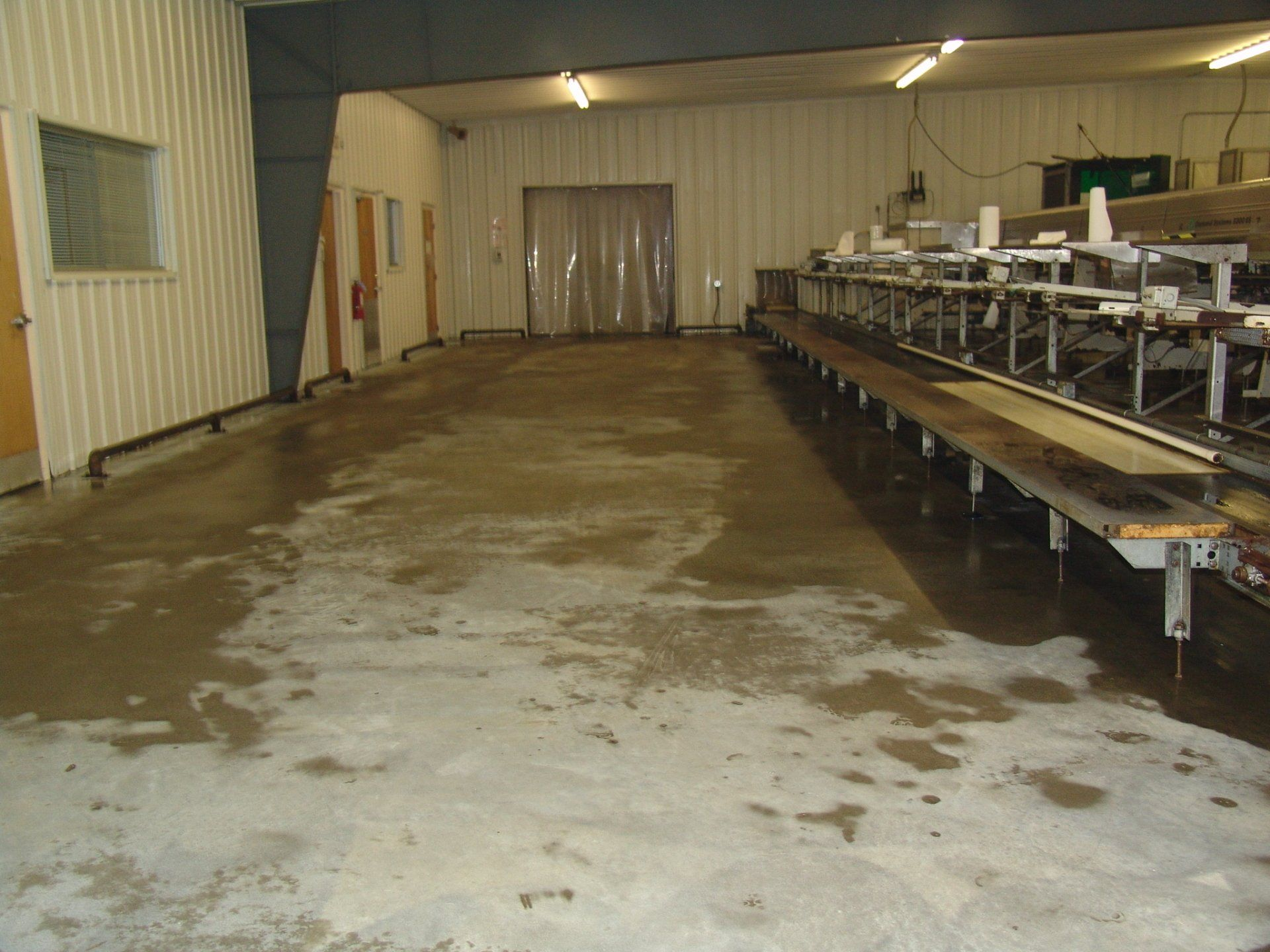 Wet concrete floor in industrial building, possibly food processing area, with equipment and plastic strip door.