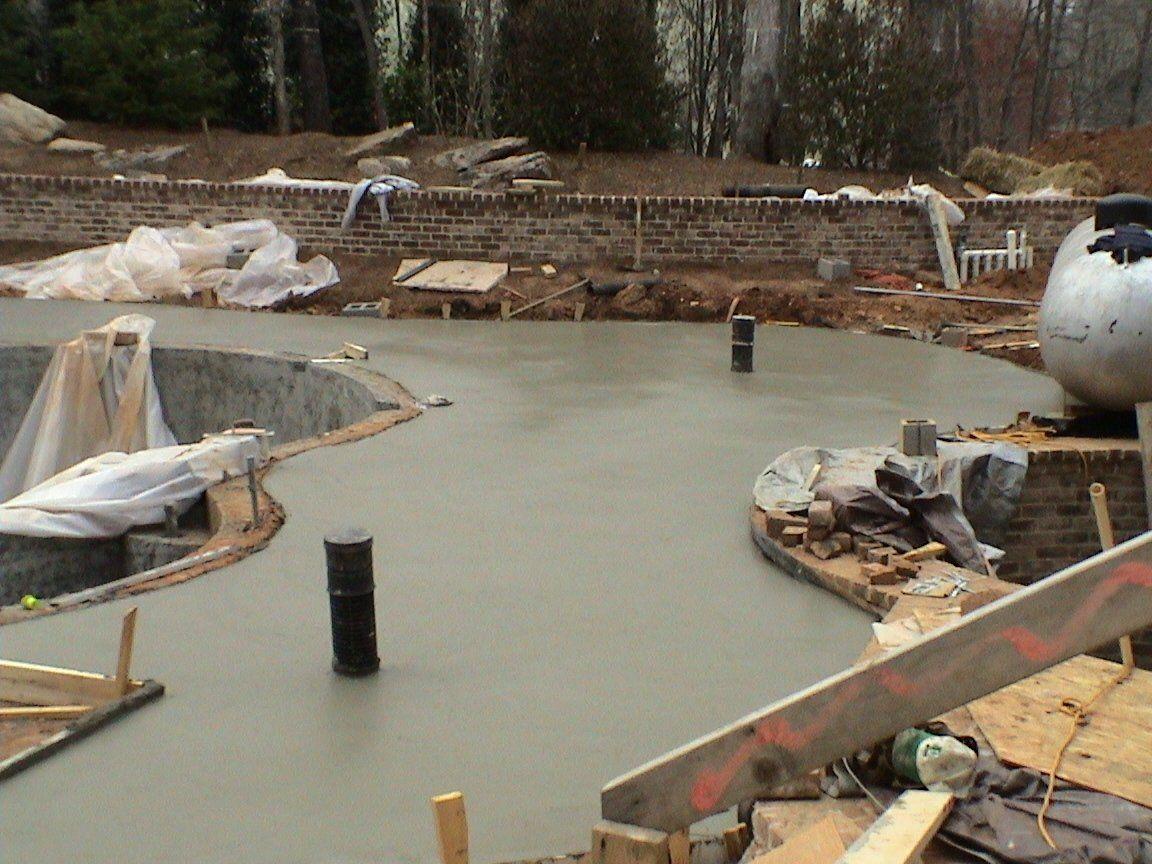 Concrete pool deck construction site with black pipes and brick retaining wall.