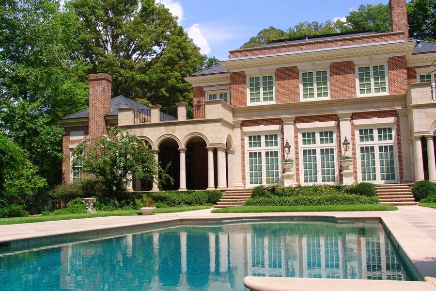 Large brick mansion with a pool, columns, and green landscaping under a bright sky.