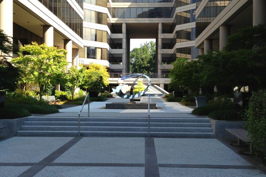 Courtyard with tiered steps, sculpture, trees, and buildings. Sunny, outdoor setting.