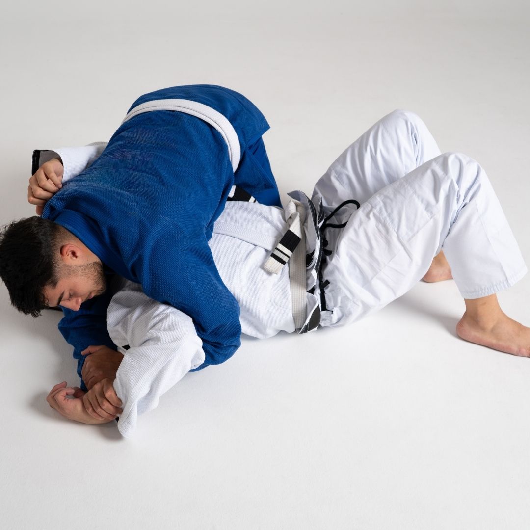 Two men in grappling position on a mat. One embraces the other from behind, both smiling. Gym setting.