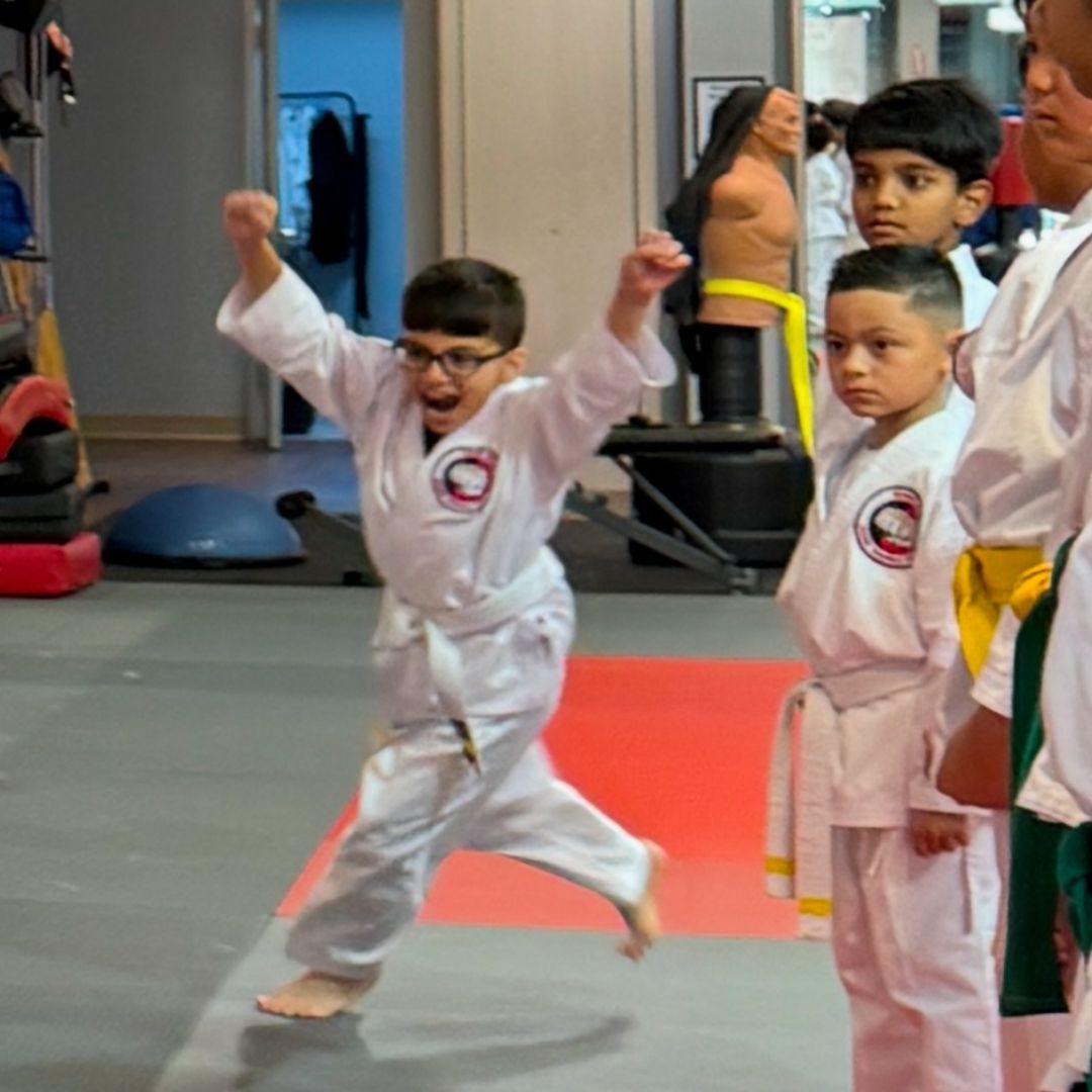 Children in colorful gis play a game on a mat. Two instructors pull a student in blue.