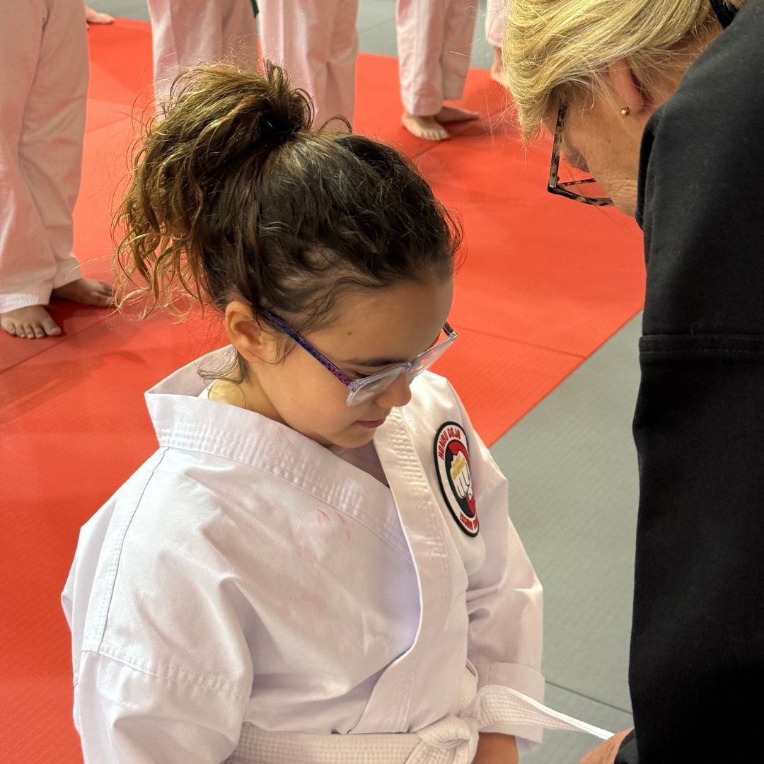 Two children practicing Brazilian Jiu-Jitsu on a mat. One child in white gi has the other in a leg lock. A girl watches.