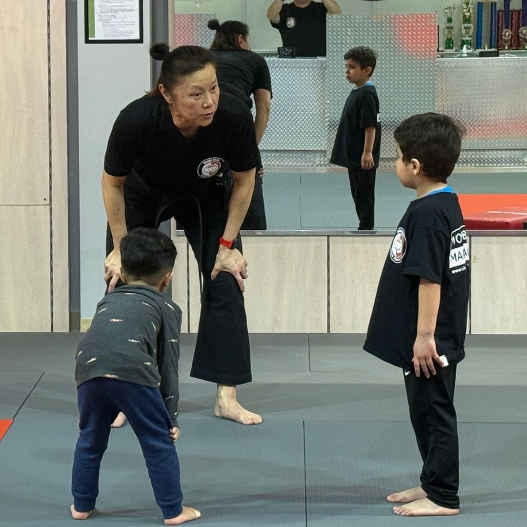 Two children in black and white martial arts uniforms practice a chokehold on a mat. One child smiles.