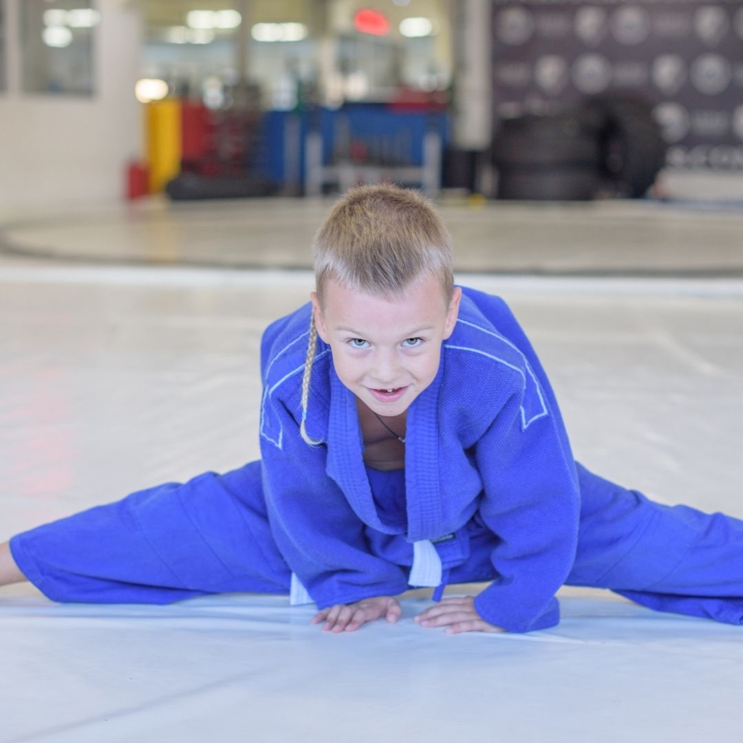 Children in colorful gis play a game on a mat. Two instructors pull a student in blue.