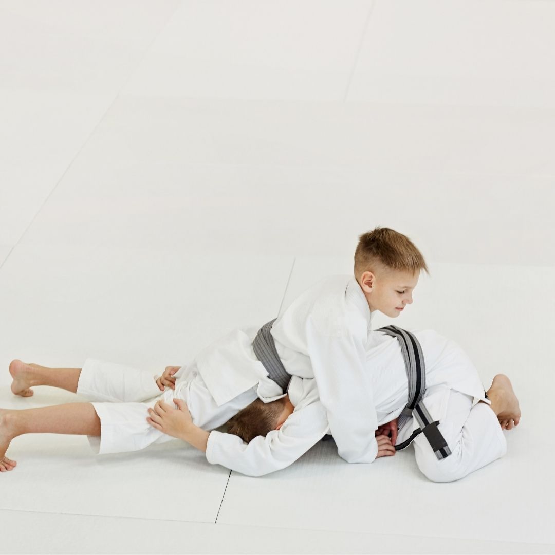 Two boys on a mat practicing Jiu-Jitsu. One boy is on top, giving a thumbs-up.