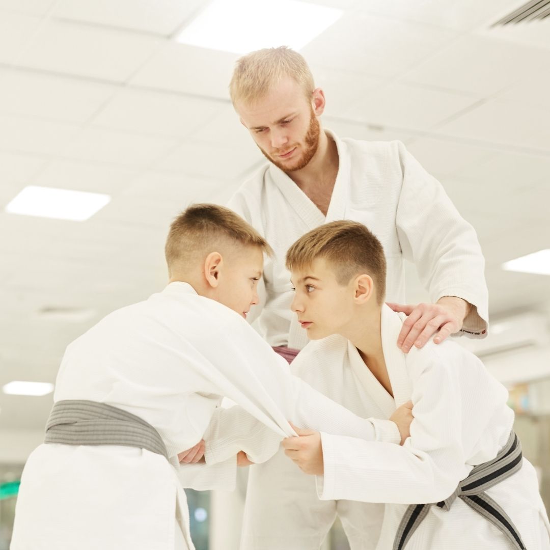 Two young children in blue and black martial arts uniforms grappling on a gym floor, one smiling.