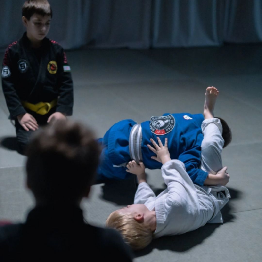 Two children practicing Brazilian Jiu-Jitsu on a mat. One child in white gi has the other in a leg lock. A girl watches.