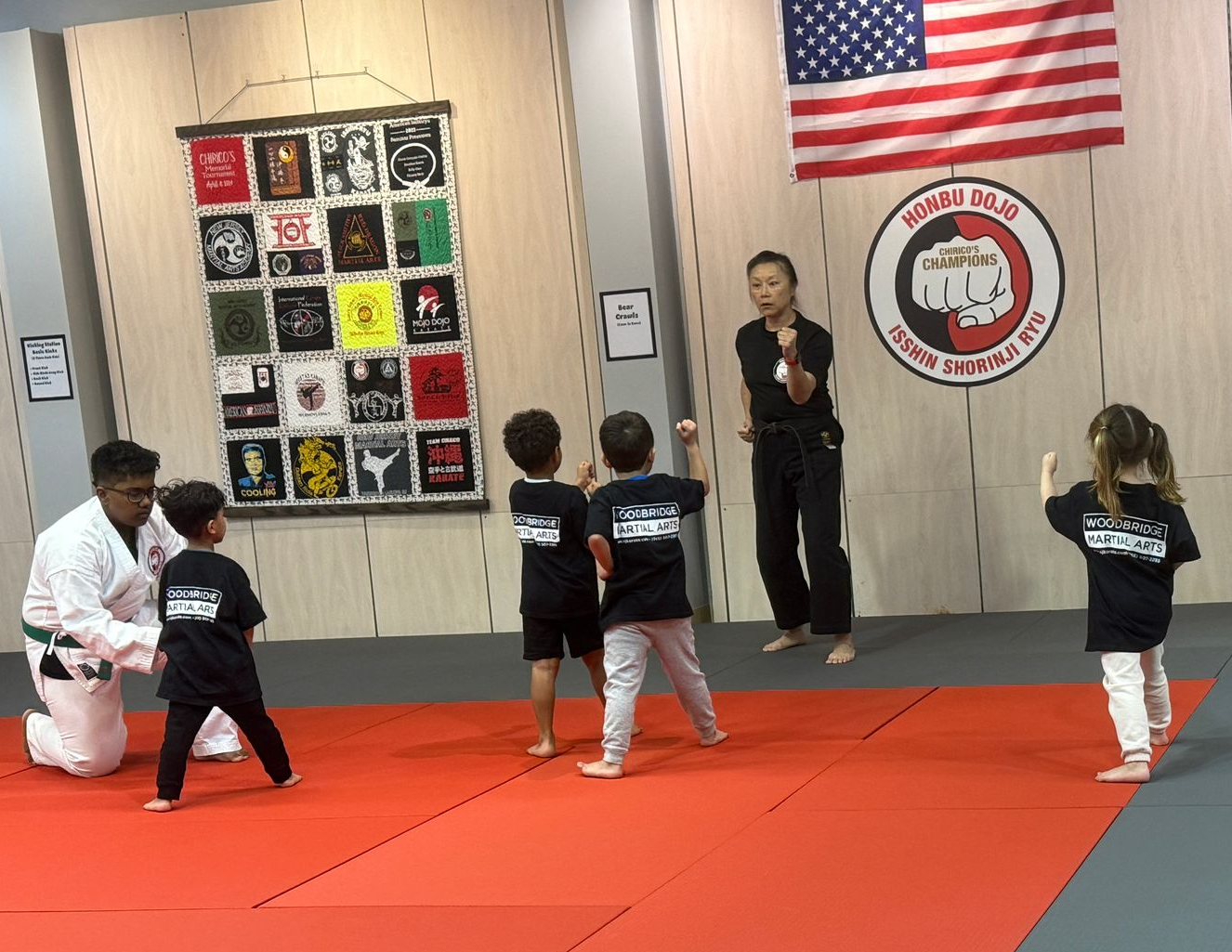 Two young children in blue and black martial arts uniforms grappling on a gym floor, one smiling.