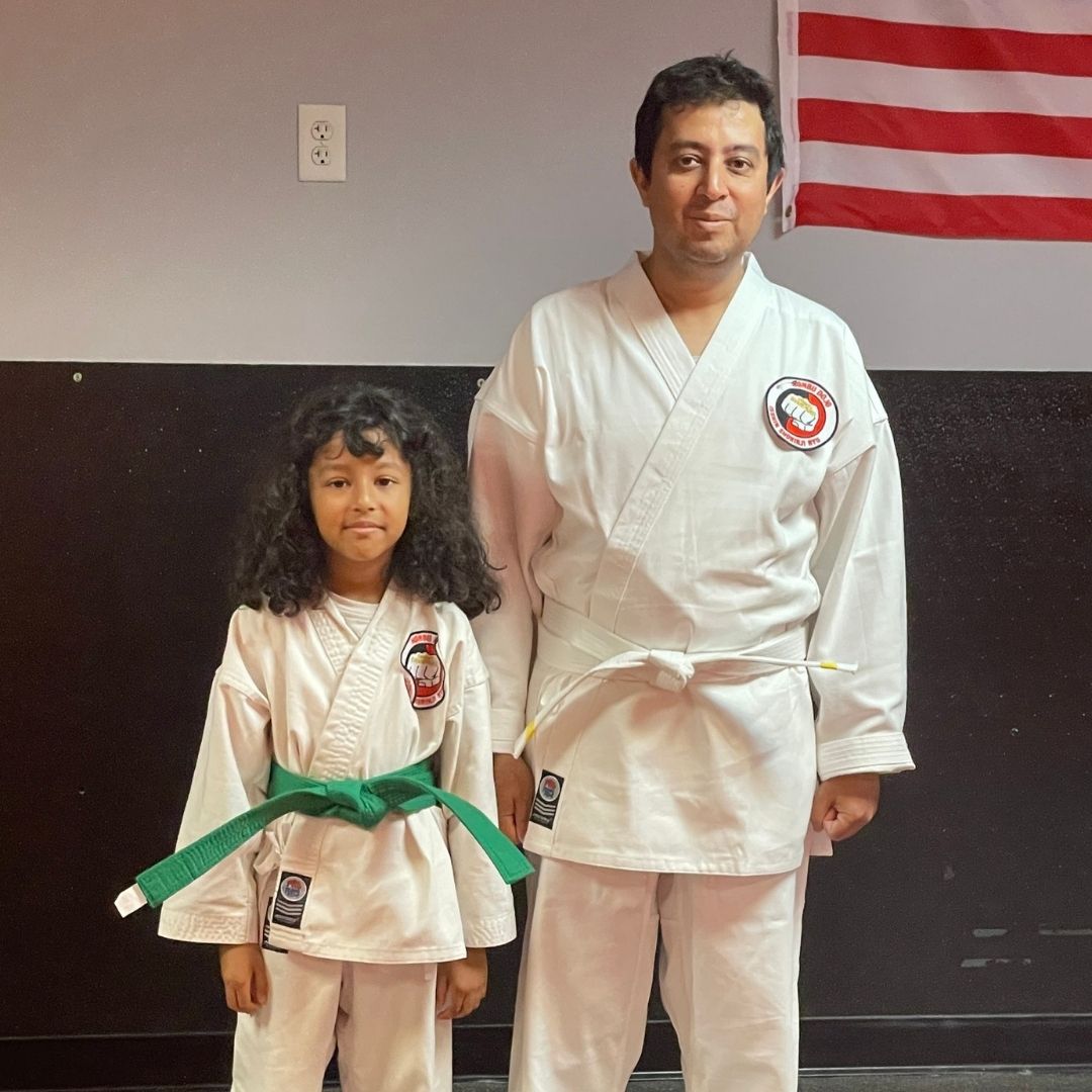 Family in Jiu-Jitsu gis poses. Indoors, blue mats, a smiling woman in the middle, around a seated man with a beard.