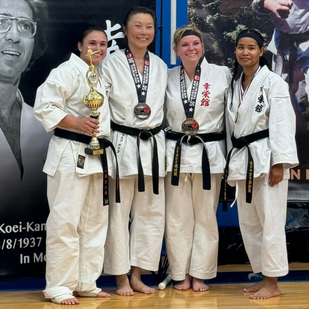 Five people in jiu-jitsu uniforms on a blue mat, including a man with a red belt, posing in a gym.