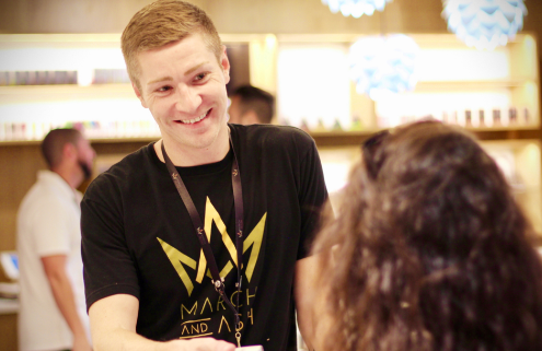 Man smiles, talks to customer in a store. He wears a black shirt with gold logo and an ID badge.
