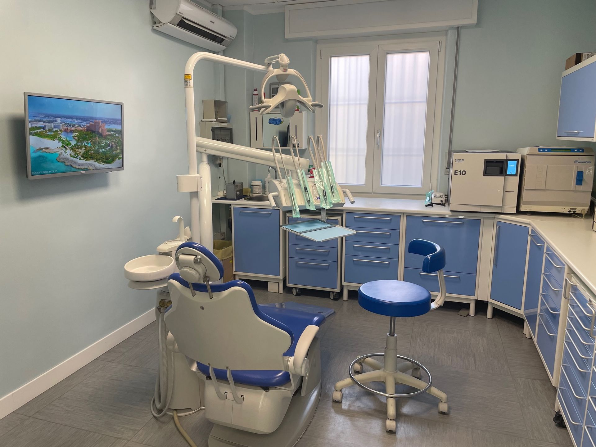 Studio dentistico con poltrona, attrezzatura e armadietti, nei toni del blu e del bianco. Sono visibili una finestra e una TV.