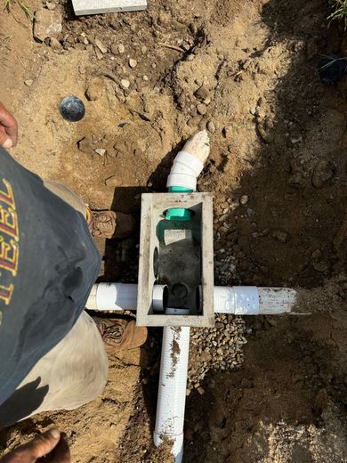 Plumbing setup in dirt with a worker's arm visible, featuring pipes and a concrete box.
