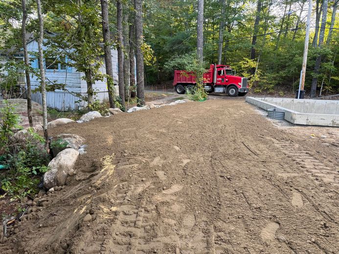 A red dump truck unloading dirt near a building and trees on a sunny day.