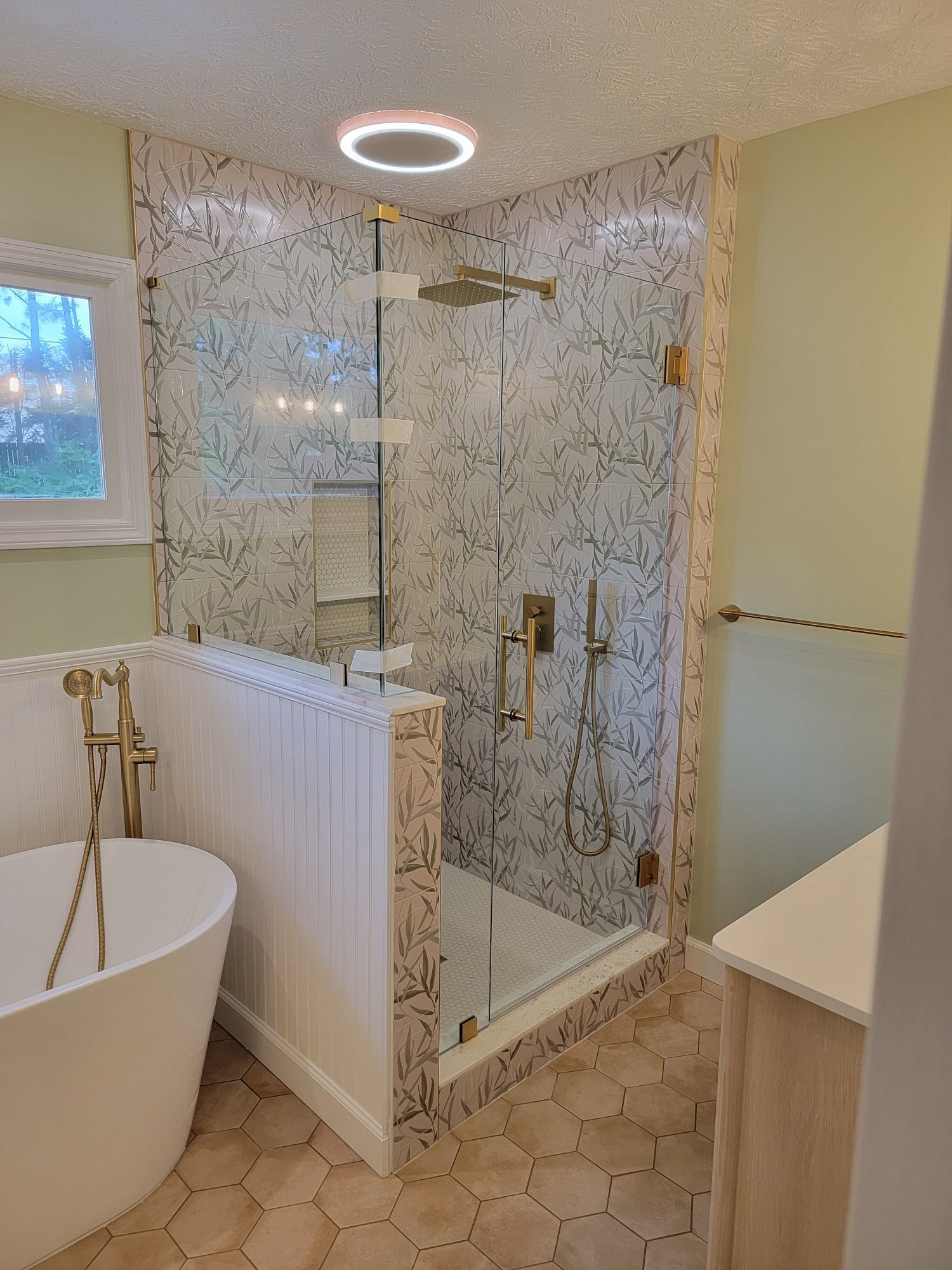 Bathroom with a glass shower, soaking tub, and gold fixtures. The walls are light green, and the floor has hexagonal tiles.