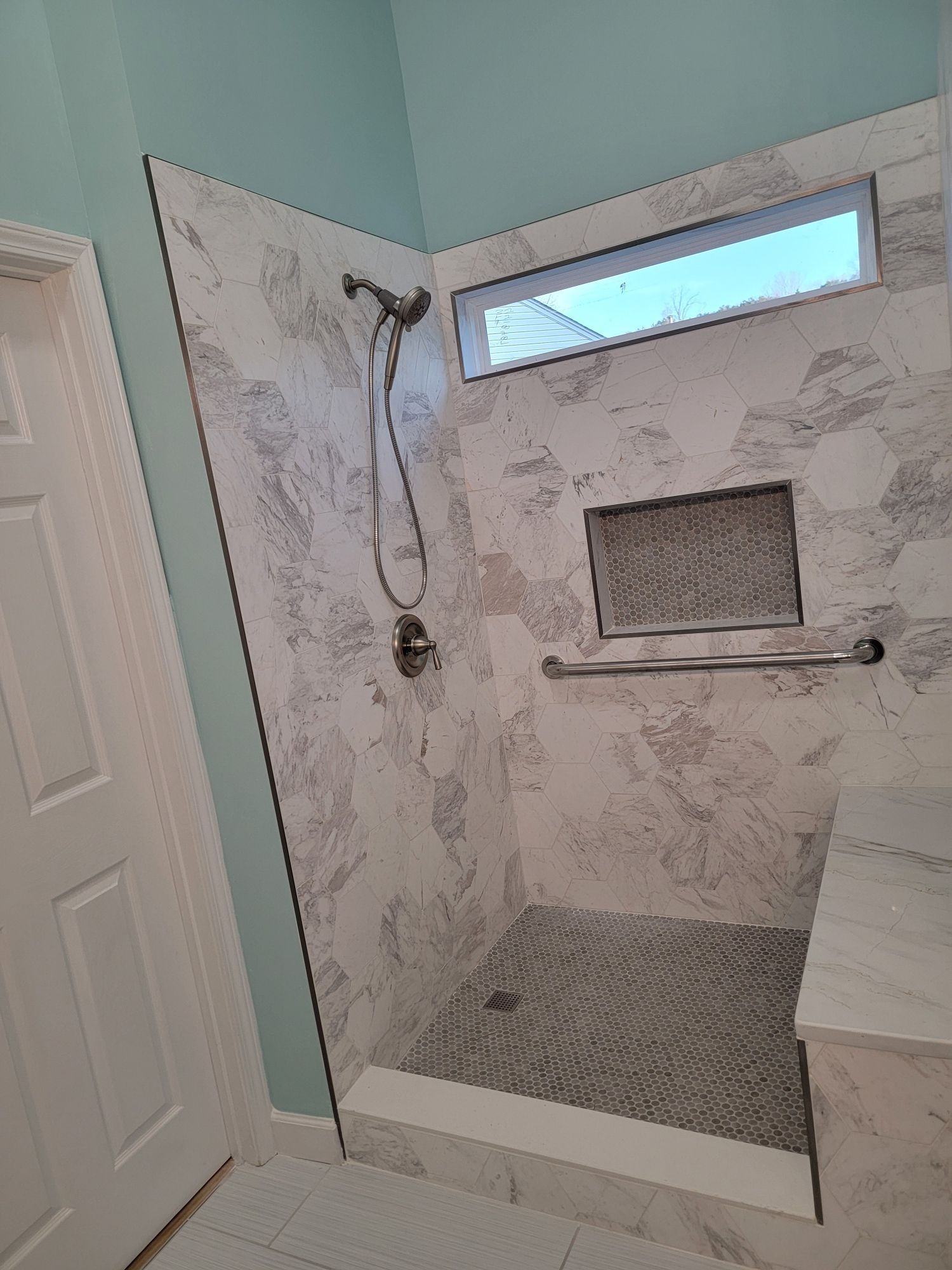 Shower stall with marble-patterned tile, glass door, window, and built-in bench. Includes a handrail and shower head.