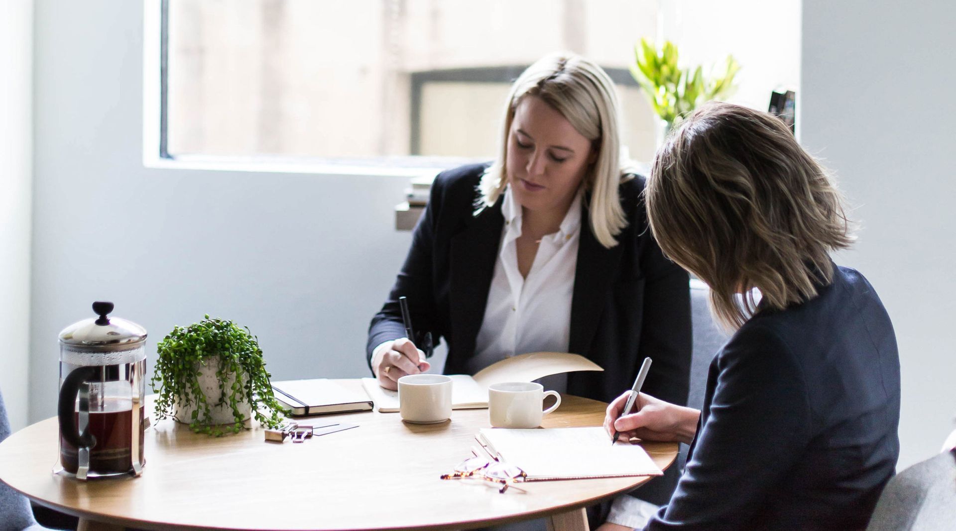 Two women in a bright office writing at a round table, coffee and plants nearby.