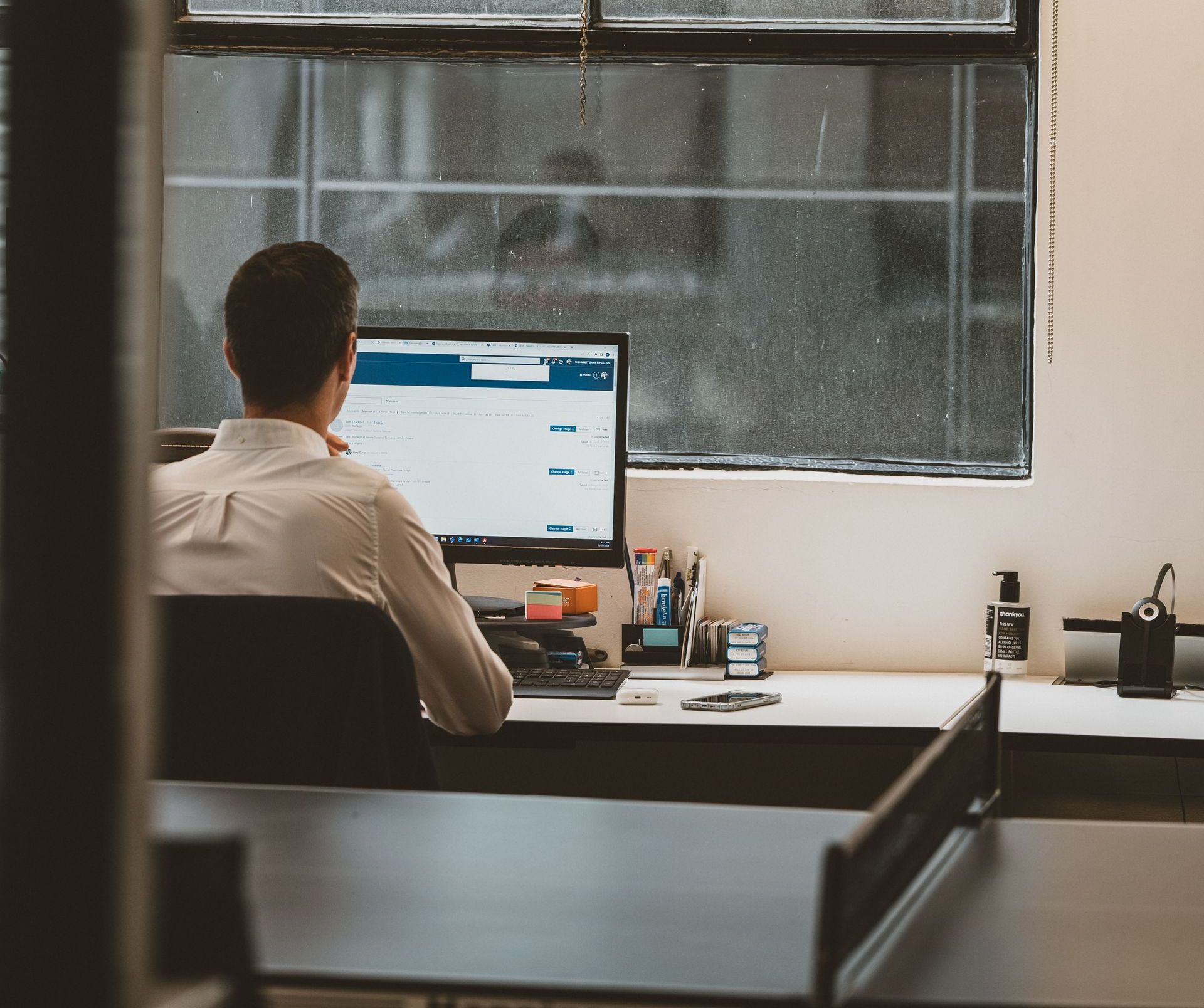 Man at computer desk, facing screen, in modern office setting near a window.