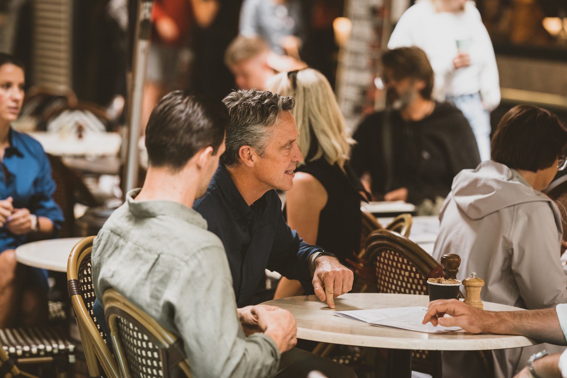People at outdoor cafe tables, conversing, pointing at papers. Sunny setting, neutral tones.