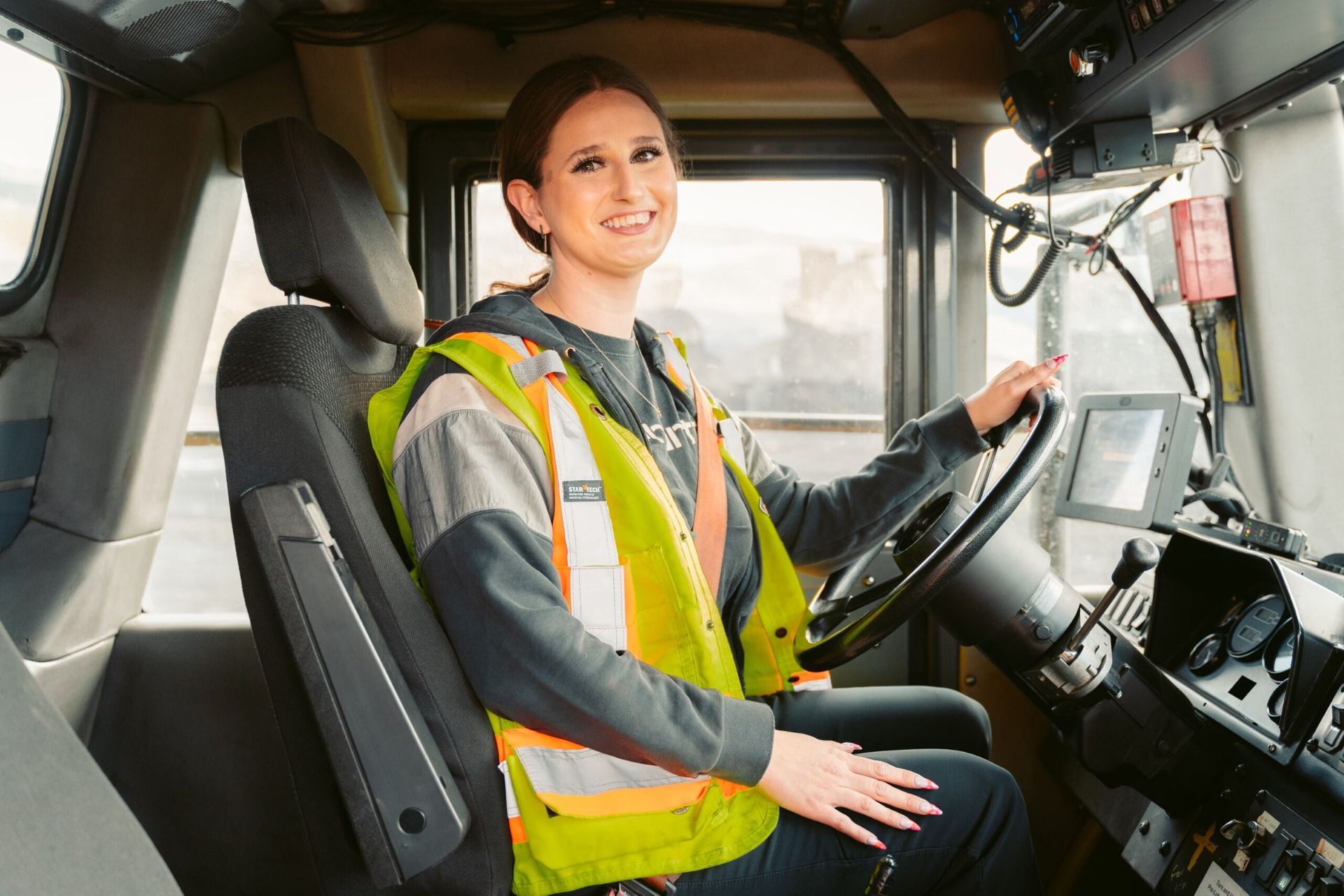 A female Haul Truck Operator wearing personal protective equipment operating heavy machinery, holding the steering wheel.