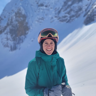 A selfie of George wearing a ski helmet with goggles and an orange jacket smiling while outside in the snow.