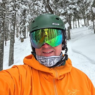 A selfie of George wearing a ski helmet with goggles and an orange jacket smiling while outside in the snow.