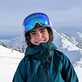 Kate, wearing a ski helmet with goggles and a green jacket is smiling while at Fernie Alpine Resort in the winter. 