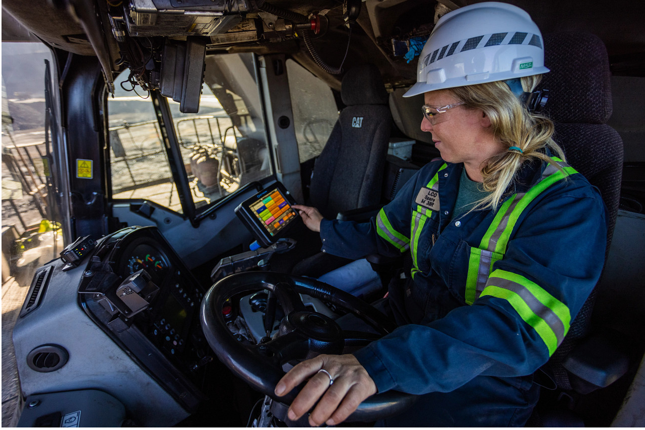 A female Haul Truck Operator wearing personal protective equipment operating heavy machinery, holding the steering wheel.