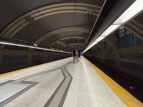 Subway platform with arched ceiling and bright overhead lights. Two people stand near a train.