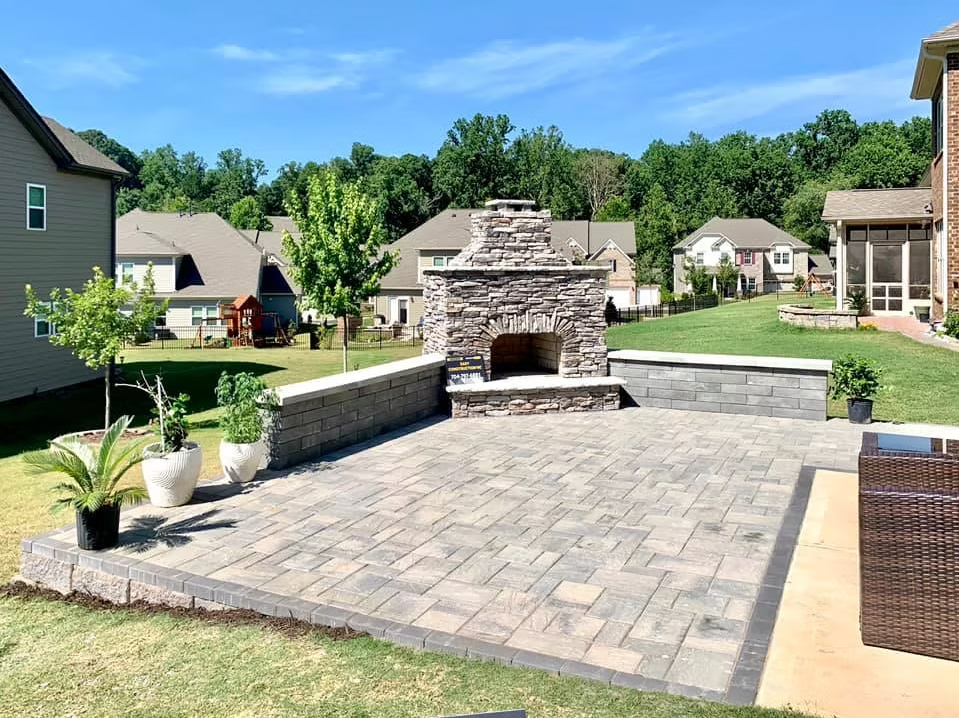 Stone patio with fireplace, built-in walls, and potted plants. Houses and trees in the background under a blue sky.