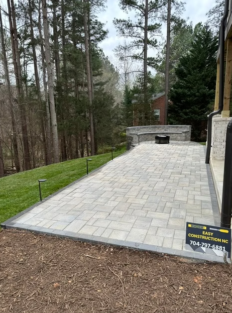 Paver patio with a stone fireplace, bordered by grass and trees.