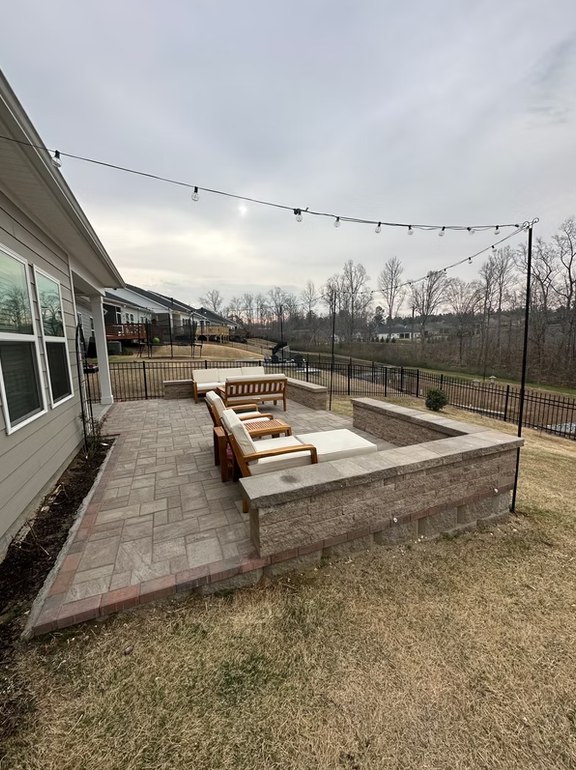 Patio with pavers, built-in seating, and a fire pit, under string lights, by a house on a cloudy day.