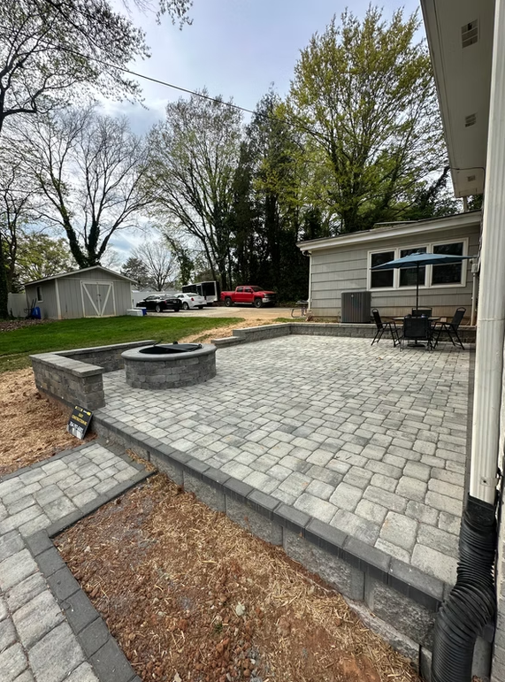 A paved patio with a fire pit, bordered by a retaining wall, next to a house under a cloudy sky.
