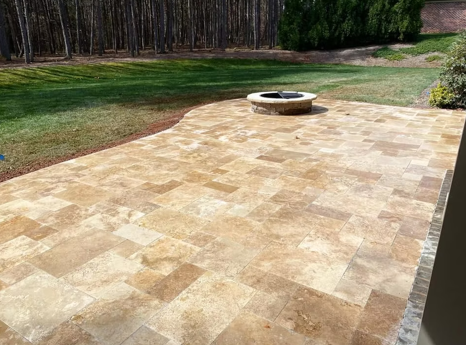 Stone patio with fire pit, bordered by grass and trees.