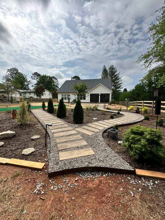 Stone paths split toward a white building with black garage doors, surrounded by landscaping, under a cloudy sky.
