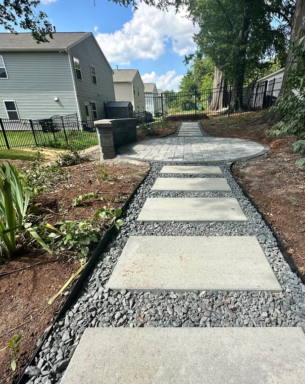 Pathway with rectangular stepping stones, surrounded by dark gray gravel and bordered by black edging, leading to a brick patio with a grill.