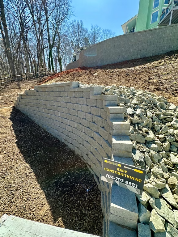 Curved retaining wall of gray blocks on a hillside, under a blue sky, with construction debris.