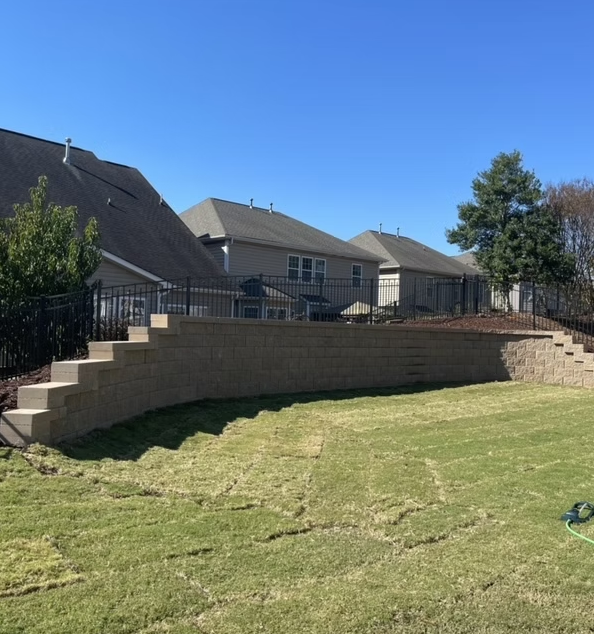 A backyard with a tiered retaining wall, steps, and black fence. Houses are in the background, blue sky overhead.
