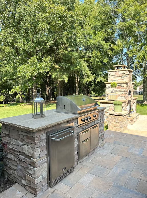 Outdoor kitchen with stainless steel grill, fridge, and pizza oven, on a stone patio with trees in the background.