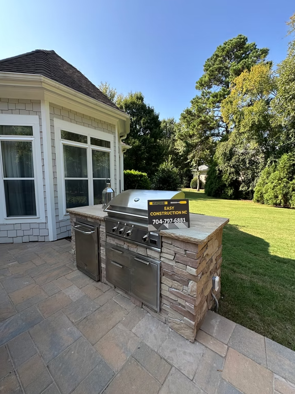 Outdoor kitchen with a stainless steel grill and stone facade, next to a house on a sunny day.