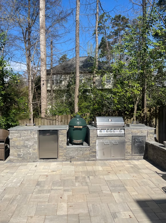 Outdoor kitchen with stone counters, a grill, Big Green Egg, and stainless steel appliances. Paved patio setting.
