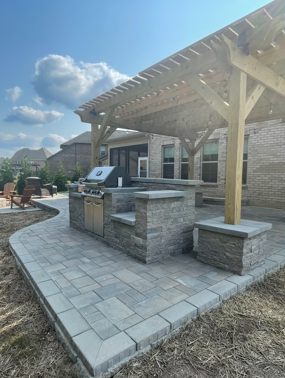 Outdoor kitchen with grill, stone countertop, and wooden pergola on a brick patio.