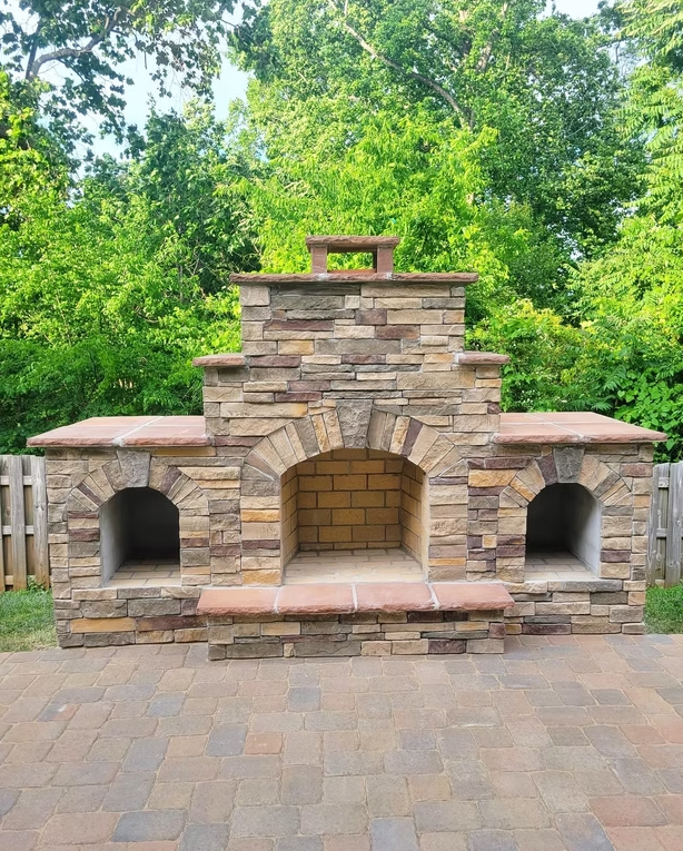 Stone outdoor fireplace with brick interior, flanked by storage spaces, set on a brick patio with trees in the background.