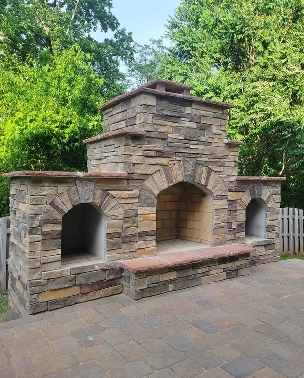 Outdoor stone fireplace with arched openings and a brick base, set against a green tree backdrop.