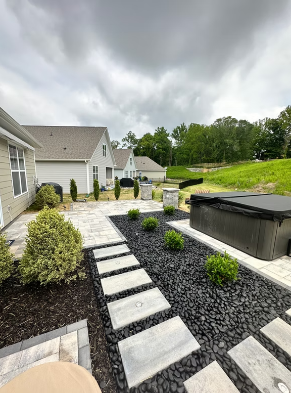 Backyard patio with hot tub, stepping stones, and dark landscaping. Houses and trees in background.