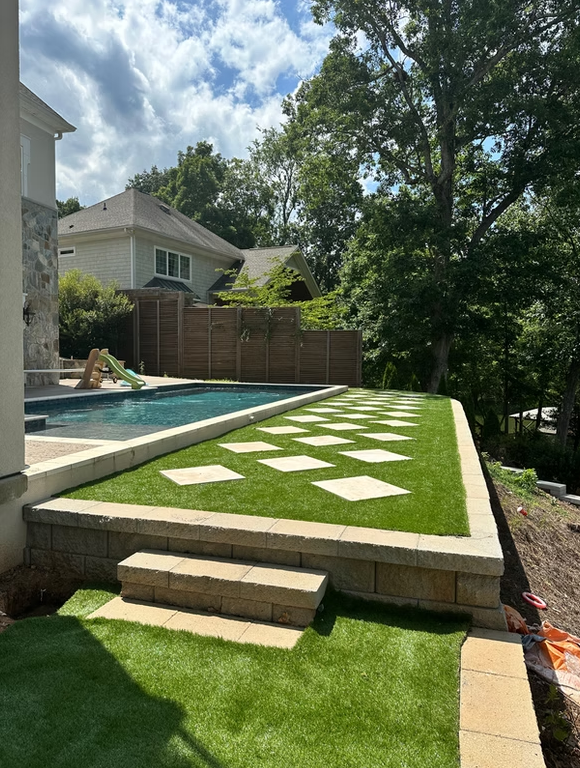 Pool and patio area with artificial turf, stone steps, and a wooden fence on a sunny day.