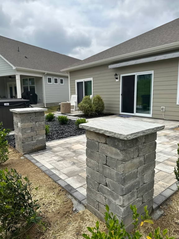 Stone columns frame a paved patio beside a house. Dark mulch beds and landscaping complete the outdoor space.