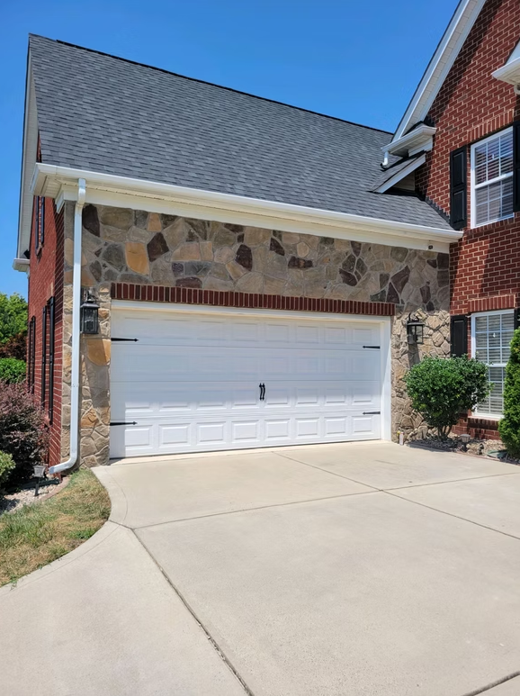 Garage with white door, stone facade, and red brick house under a blue sky.