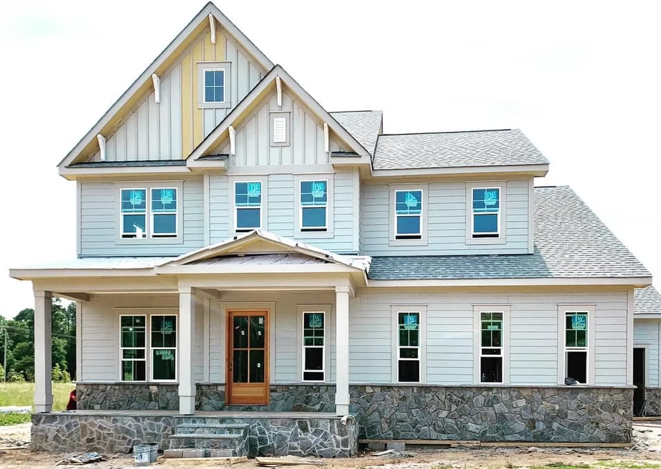 Two-story house under construction with light blue siding, stone base, and wooden door.